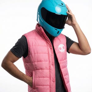 Puede incluir: Un casco de moto turquesa brillante con una visera negra. La persona lleva un chaleco acolchado rosa con un gráfico de calavera blanca. El chaleco tiene una parte delantera abotonada y bolsillos laterales. La persona lleva una camiseta gris oscuro.