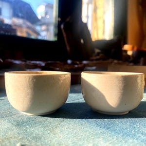 May include: Two off-white ceramic bowls with a rounded shape and a textured surface. The bowls are sitting on a blue surface, with sunlight casting shadows across them. The bowls are likely for serving food or beverages.
