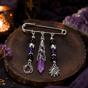 Peut inclure: Une broche épingle de sûreté argentée avec trois breloques pendantes. La breloque centrale est un cristal violet pointu dans une coiffe en filigrane. Les breloques latérales se composent de perles noires et violettes avec un croissant de lune.