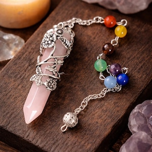 May include: A rose quartz pendulum with a silver-colored floral design and a chain of colorful beads. The pendulum is on a wooden surface, with a lit candle and crystals in the background. The beads are red, yellow, brown, green, purple, blue.