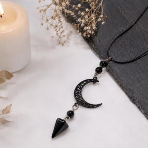 Peut inclure: Un collier noir composé d'un pendentif en forme de croissant de lune avec des découpes en filigrane, suspendu à un cordon noir. Le design comprend deux petites perles noires sphériques au-dessus de la lune et une pierre noire pointue.