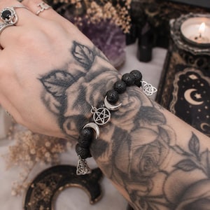 Peut inclure: Bracelet de perles en pierre de lave noire avec des breloques argentées. Les breloques comprennent un pentagramme, des croissants de lune et un nœud celtique. Un accessoire sombre et terreux sur le thème mystique.