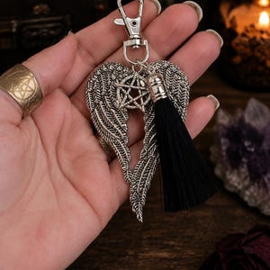 Peut inclure: Porte-clés argenté avec des ailes d'ange, un charme pentagramme et un pompon noir. Les ailes sont détaillées avec des textures en forme de plumes. Le porte-clés a un fermoir mousqueton pour une fixation facile.