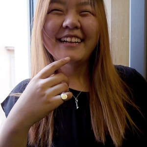 Puede incluir: Una joven con el pelo largo y castaño sonríe y se señala la barbilla. Lleva una camisa negra y un collar de plata con un colgante de llave. También lleva un anillo de plata en el dedo.