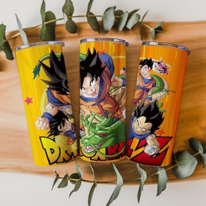 Op de afbeelding: Drie gele en oranje geïsoleerde bekers met Dragon Ball Z-personages. Elke beker heeft een ander karakterontwerp, met levendige kleuren en gedetailleerde illustraties. De bekers zijn cilindrisch en hebben een zilveren deksel.
