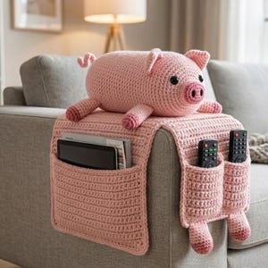 May include: A light pink crocheted sofa armrest organizer with pockets for remotes and a tablet. A matching pink crocheted pig sits on top. The organizer is designed to fit over the arm of a sofa.