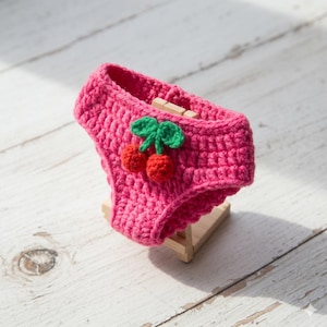 Può includere: Un pannolino per animali domestici all'uncinetto rosa acceso con un motivo a ciliegia. Il pannolino presenta una ciliegia rossa con foglie verdi. L'accessorio per animali fatto a mano è esposto su un piccolo supporto di legno.