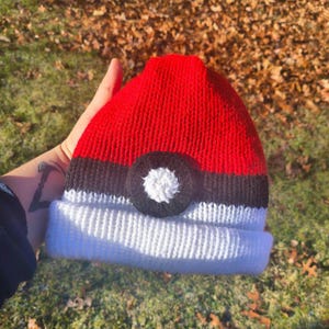 May include: A red, black, and white knit beanie hat. The hat features a red top, a black band, and a white brim. A black circle with a white center is on the front of the hat. The hat is made of a soft, knitted material.