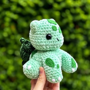 May include: A light green crocheted plush toy with dark green leaf accents and white spots. The toy has a round head, black eyes, and small arms and legs. The background is a blurred green.