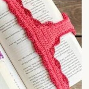 May include: A handmade, coral-colored crochet bookmark rests on the open pages of a book. The bookmark has a scalloped edge and is made of a soft, textured yarn. The book is open to a page of printed text.