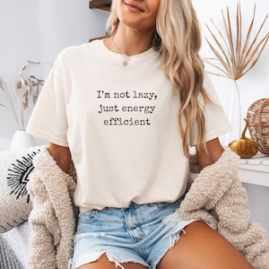Puede incluir: Camiseta color crema con el texto negro "I'm not lazy, just energy efficient". La camiseta se combina con pantalones cortos vaqueros y una rebeca beige. El conjunto transmite un estilo casual.