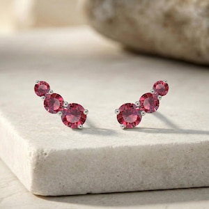 Peut inclure: Une paire de boucles d'oreilles grimpantes argentées. Chaque boucle d'oreille présente trois pierres précieuses rondes, rouge vif, de taille progressive. Les pierres sont serties dans du métal argenté, créant un design délicat et élégant.