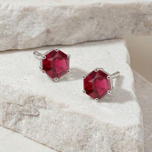 May include: A pair of silver cufflinks with large, faceted, hexagonal, ruby-red gemstones. The cufflinks are set against a light-colored, textured background, showcasing their vibrant color and design. The gemstones are approximately 0.75 inches in diameter.