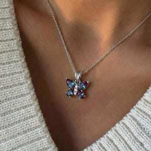 May include: A silver necklace with a butterfly pendant. The pendant is adorned with purple and teal gemstones and small white stones. The butterfly is suspended from a delicate silver chain. The necklace is displayed on a person wearing a white sweater.