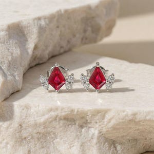 May include: A pair of silver stud earrings with a vibrant red, diamond-shaped center stone. Each earring features a cluster of small, clear stones on either side of the red stone. The earrings are displayed on a light-colored, textured surface.