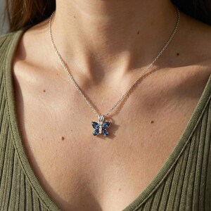 May include: A silver necklace with a butterfly pendant. The pendant has a butterfly design with blue gemstones and a small diamond in the centre. The necklace is worn on a person.