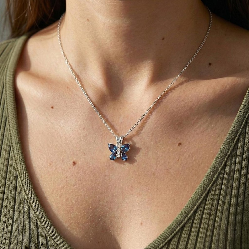 May include: A silver necklace with a butterfly pendant. The pendant has a butterfly design with blue gemstones and a small diamond in the centre. The necklace is worn on a person.