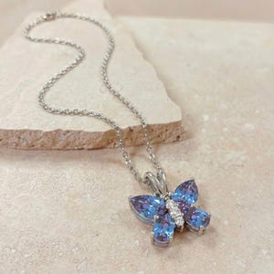 May include: A silver necklace with a butterfly pendant. The pendant has blue and purple gemstones and a cluster of small white gemstones. The necklace is displayed on a light-coloured surface.