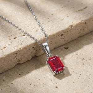 Könnte beinhalten: Eine silberne Halskette mit einem rechteckigen, tiefroten Edelsteinanhänger. Der Anhänger ist in einen silbernen Rahmen gefasst und hängt an einer filigranen Silberkette. Die Halskette wird auf einer strukturierten, hellfarbigen Oberfläche präsentiert.