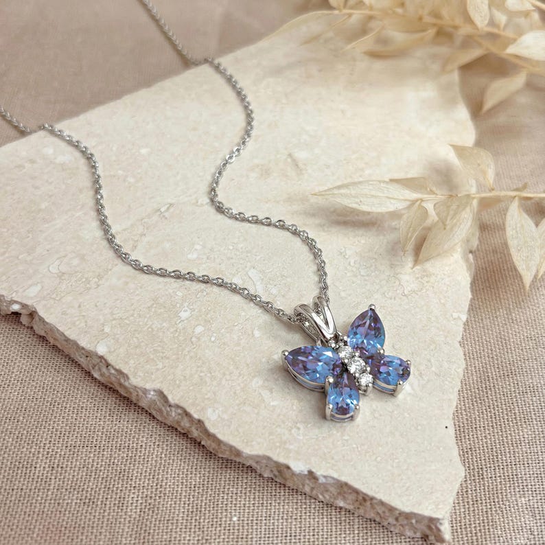 May include: A silver butterfly pendant necklace with blue and white gemstones. The butterfly design features a cluster of blue teardrop-shaped stones and small white stones. The necklace chain is delicate and silver.