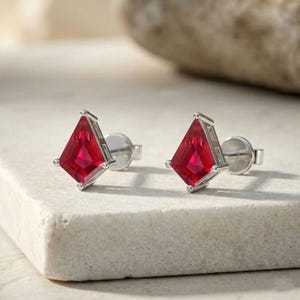 May include: A pair of silver stud earrings with kite-shaped, vibrant red gemstones. The earrings have a geometric design with a polished finish, set against a neutral background. The gemstones are approximately 0.5 inches in length.