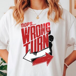 May include: A white short-sleeved t-shirt featuring a graphic print with the text OOPS... WRONG TURN in distressed red lettering. The design includes a black silhouette of a grinning figure and two directional arrows, one white and one red.