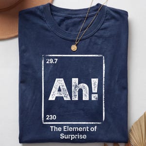 May include: A navy blue t-shirt featuring a white graphic designed like a periodic table element box. The graphic displays the text 29.7, Ah!, 230, and the phrase The Element of Surprise. The shirt has a crew neckline and short sleeves.