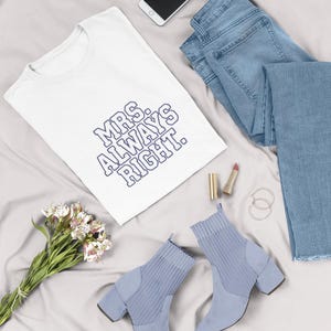 May include: A flat lay featuring a white t-shirt with the text "MRS. ALWAYS RIGHT" in blue, light blue jeans, a pair of light blue ankle boots, a smartphone, lipstick, and a bouquet of flowers on a gray surface.