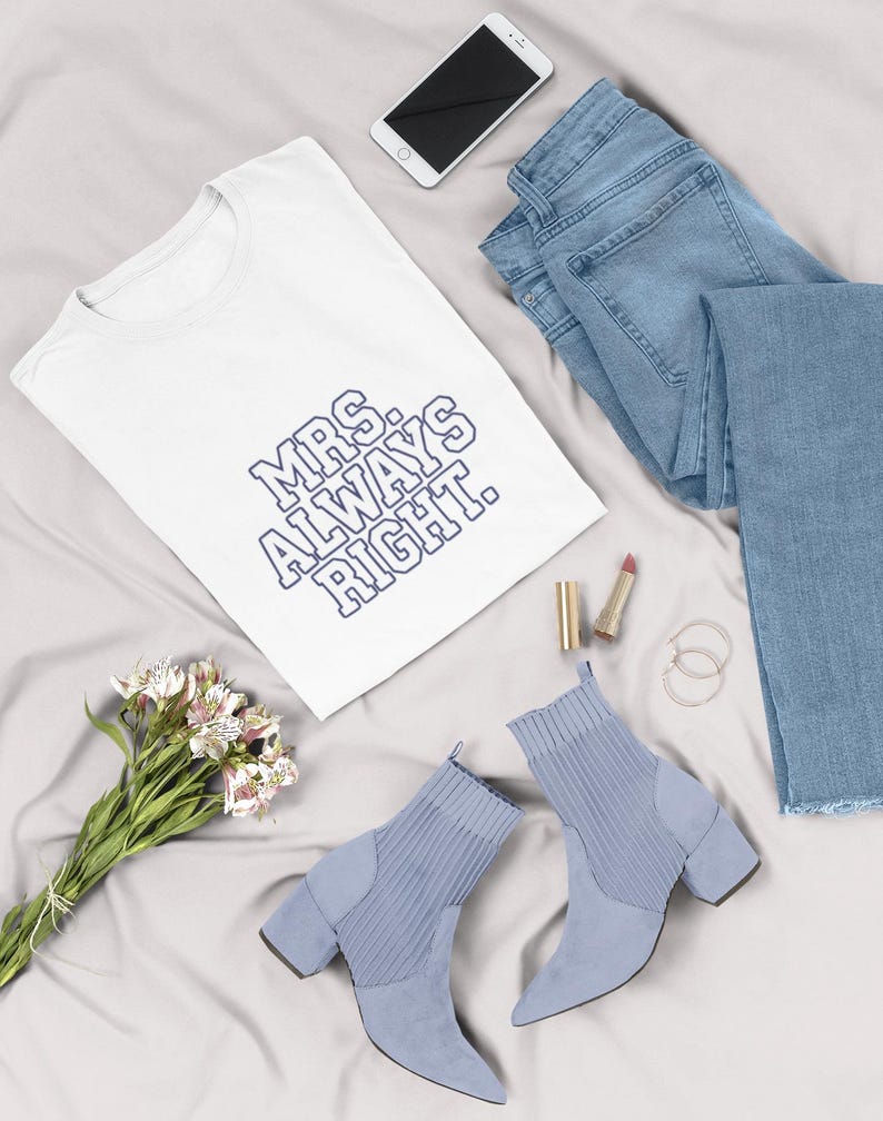 May include: A flat lay featuring a white t-shirt with the text "MRS. ALWAYS RIGHT" in blue, light blue jeans, a pair of light blue ankle boots, a smartphone, lipstick, and a bouquet of flowers on a gray surface.