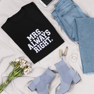 May include: A black t-shirt with the text "MRS. ALWAYS RIGHT" in white, paired with light blue jeans, a smartphone, and light blue heeled ankle boots. A bouquet of white and pink flowers and lipstick complete the ensemble.