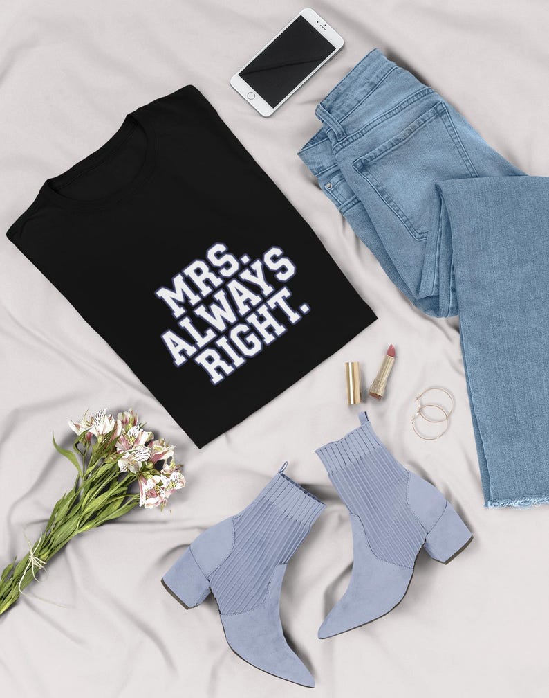 May include: A black t-shirt with the text "MRS. ALWAYS RIGHT" in white, paired with light blue jeans, a smartphone, and light blue heeled ankle boots. A bouquet of white and pink flowers and lipstick complete the ensemble.