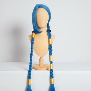May include: A blue wig with long, braided pigtails. The wig is displayed on a wooden mannequin head. The braids are accented with gold-colored bands. The wig has a short, angled front and is a vibrant shade of blue.