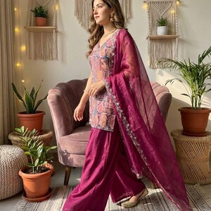 May include: A magenta salwar kameez outfit. The top has a V-neck and detailed embroidery, paired with wide-leg trousers. A matching sheer dupatta is draped. Gold embroidered flats complete the look. The background includes plants and macrame wall hangings.