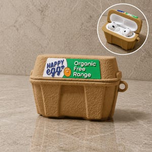 Könnte beinhalten: Eine braune Hülle für kabellose Ohrhörer, die einem Eierkarton nachempfunden ist. Auf der Hülle steht "HAPPY egg" und "Organic Free Range". Das Bild zeigt die Hülle geschlossen und geöffnet, wobei sich weiße Ohrhörer darin befinden.