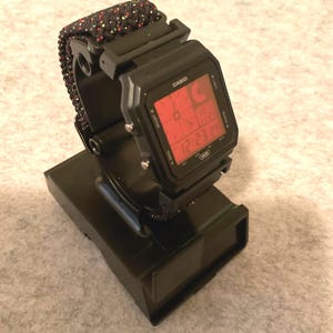 May include: A black Casio digital watch with a red display, showing the time 12:23:24, rests on a black stand. The watch has a black and red patterned band. The watch is displayed on a light gray surface.