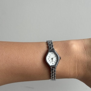 May include: A silver wristwatch with a white face and black numerals. The watch has a hexagonal face and a linked bracelet. The watch is on a wrist.
