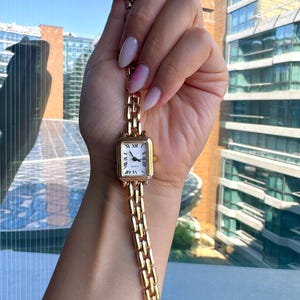 May include: A gold-coloured rectangular wristwatch with a white face and Roman numeral hour markers. The watch has a matching gold link bracelet. The watch is held up against a background of a building with glass windows.