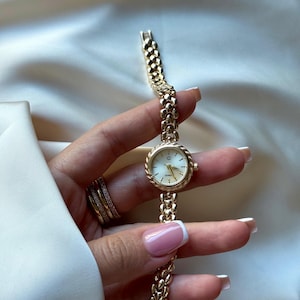 May include: A gold-toned wristwatch with a white face and gold hands. The watch has a textured bezel and a linked band. The watch is held against a white background, showcasing the watch's design and details.