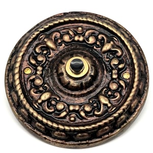 Könnte beinhalten: Verzierter Türklingelknopf im antiken Stil mit Bronze-Finish. Der kreisförmige Knopf weist detaillierte Reliefmuster und einen zentralen schwarzen Knopf auf, der von einem goldfarbenen Ring umgeben ist. Zwei kleine Schrauben befestigen den Knopf an einer Oberfläche.