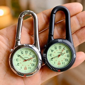 May include: Two carabiner clip watches with round faces and luminous dials. One watch has a polished silver-tone metal finish, and the other has a matte black finish. Both feature analog quartz movements, Arabic numerals, and a rotating bezel with minute markers.