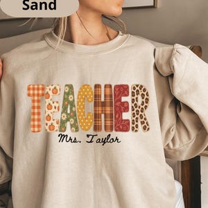 May include: A sand-coloured crewneck sweatshirt featuring the word TEACHER in large, multi-patterned letters including orange plaid, pumpkins, floral, yellow polka dots, brown plaid, red floral, and leopard print, with Mrs. Taylor written below.