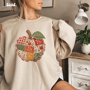 May include: A sand-colored crewneck sweatshirt featuring a large, central apple graphic composed of various quilted fabric patches. The apple design includes red gingham, floral, and polka dot patterns with a green leaf and brown stem.