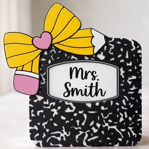 May include: A decorative teacher-themed plaque featuring a pencil and notebook design. The plaque has a yellow pencil bow with a pink heart, and the notebook has the name "Mrs. Smith" on it. The background is black and white.