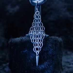 Puede incluir: Un pendiente individual de cota de malla plateada con un colgante en forma de cono. El pendiente está hecho de pequeños anillos metálicos entrelazados, formando un diseño de diamante. Cuelga de un gancho y se muestra sobre un fondo oscuro y texturizado.
