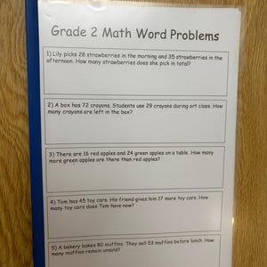 Puede incluir: Una hoja de papel blanca titulada Grade 2 Math Word Problems, dentro de una funda de plástico transparente con un lomo azul. El documento contiene cinco problemas matemáticos numerados sobre fresas, crayones, manzanas, coches de juguete y magdalenas.
