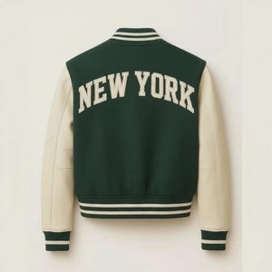 May include: A green and cream varsity jacket with the text "NEW YORK" arched across the back. The jacket features cream-colored sleeves and striped detailing on the cuffs, collar, and hem. A classic, sporty design.