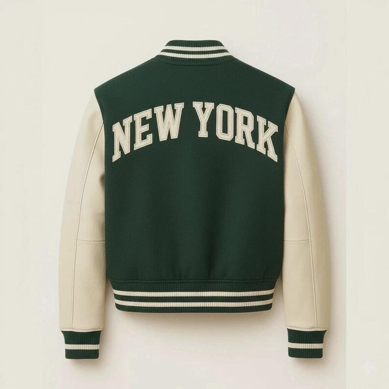 May include: A green and cream varsity jacket with the text "NEW YORK" arched across the back. The jacket features cream-colored sleeves and striped detailing on the cuffs, collar, and hem. A classic, sporty design.