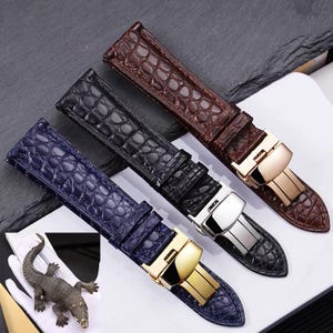 May include: Three watch straps in brown, black, and navy blue, featuring a crocodile skin texture. Each strap has a metal clasp in gold or silver. A small crocodile figurine is in the lower left corner.