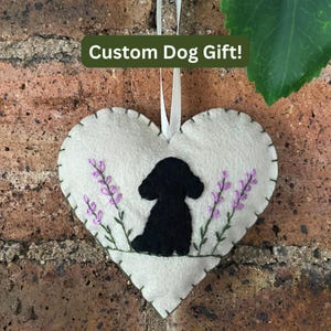 May include: A heart-shaped felt ornament in light beige featuring a black embroidered silhouette of a dog sitting between two stems of purple lavender flowers. The ornament has visible green blanket stitching along the edges and a white ribbon.