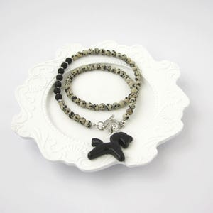Puede incluir: Un collar con un colgante de caballo negro y un diseño de cuentas. El collar tiene un cierre de palanca y se muestra en un plato blanco decorativo. Las cuentas son una mezcla de negro y blanco moteado.